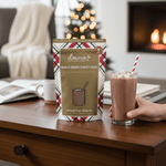 Boozy Frozen Hot Cocoa slush mix pouch with glass of frozen cocoa by fireplace and Christmas tree.