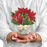 Hands holding a mini pop-up card with red poinsettias, pinecones, and greenery in a birch vase