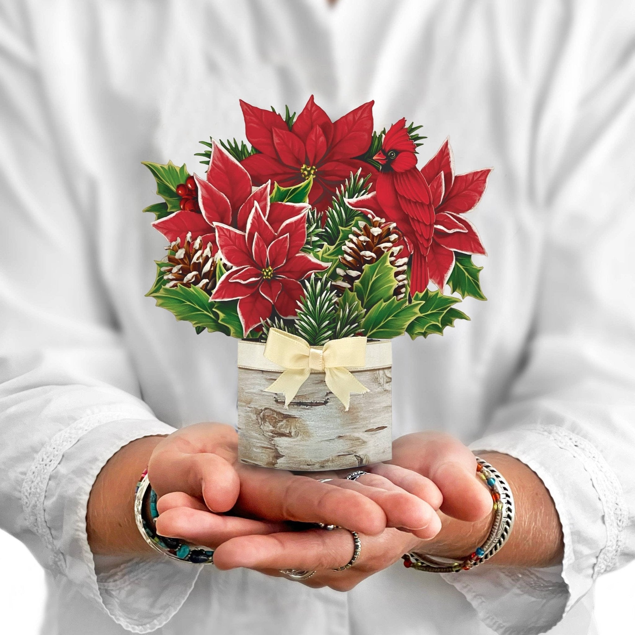Hands holding a mini pop-up card with red poinsettias, pinecones, and greenery in a birch vase