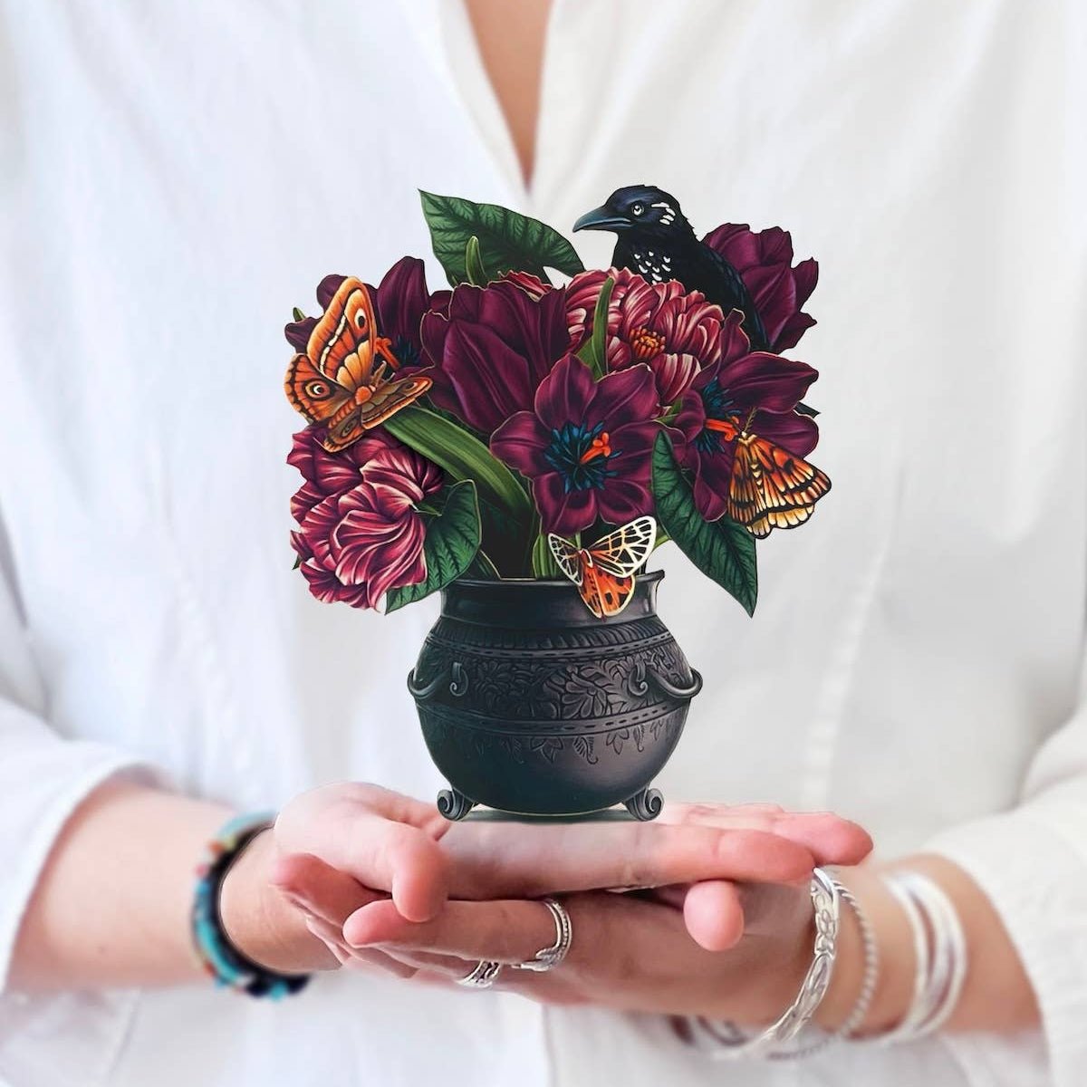 Person holding a mini pop-up bouquet with deep purple flowers, butterflies, and a raven in a black cauldron