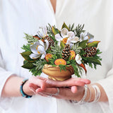 Hands holding a mini pop-up bouquet of white magnolias, pinecones, and evergreens in a wood-look vase