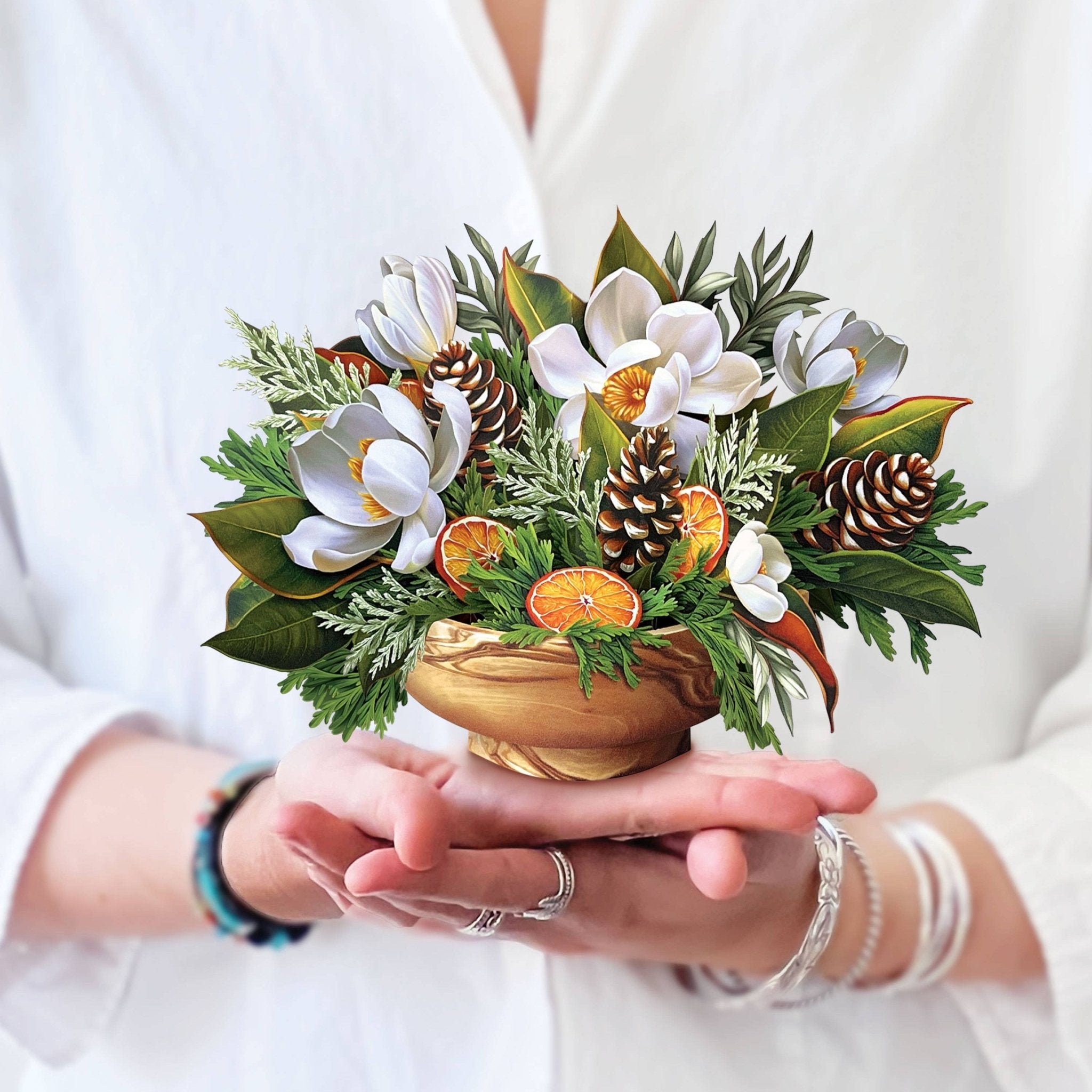 Hands holding a mini pop-up bouquet of white magnolias, pinecones, and evergreens in a wood-look vase