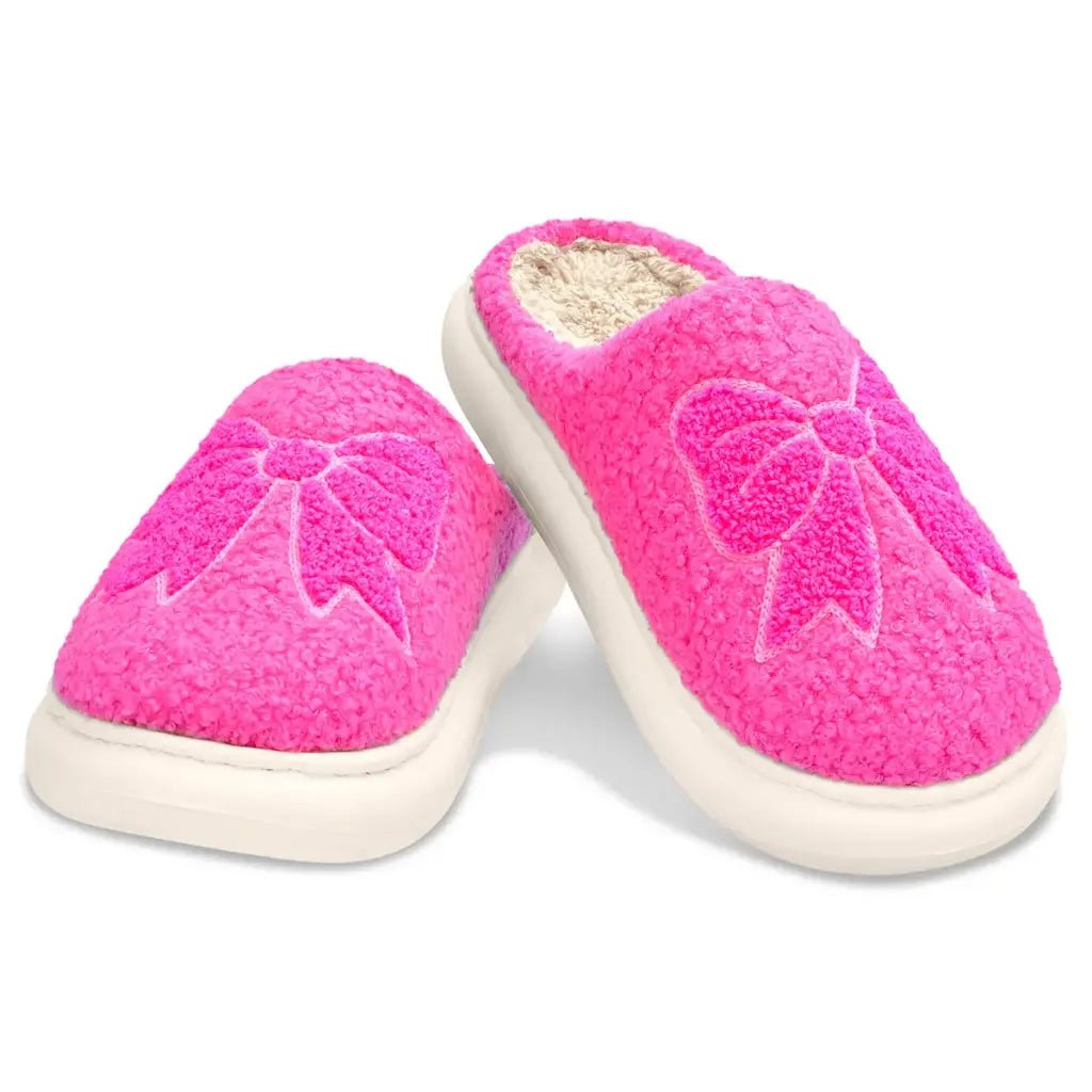 Pair of bright pink chenille slippers with embroidered bow designs and skid-resistant white soles, made for children.