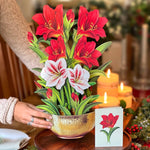 Decorative arrangement of red and white flowers in a gold pot on a wooden table with candles and Christmas decorations.