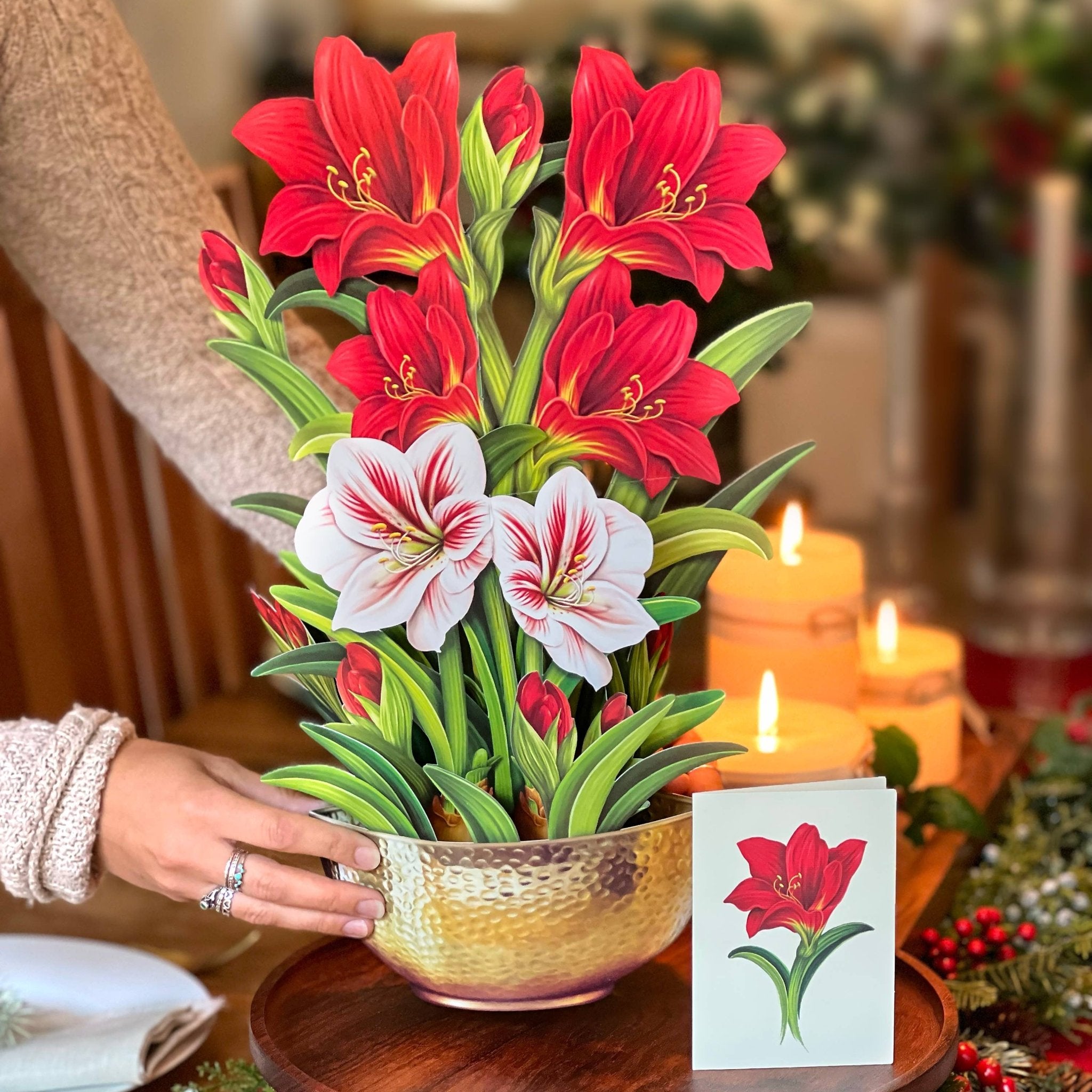 Decorative arrangement of red and white flowers in a gold pot on a wooden table with candles and Christmas decorations.