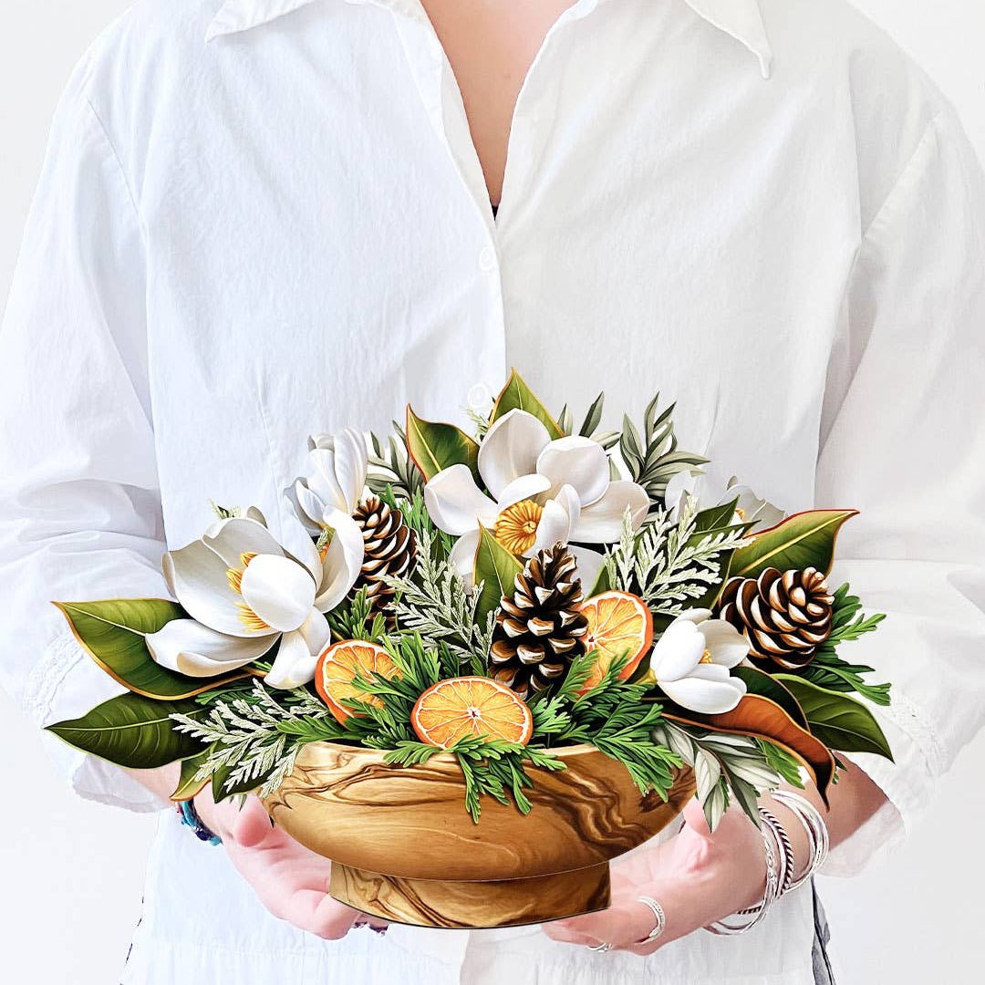 Woman holding a Winter Magnolia pop-up bouquet featuring magnolias, pine cones, oranges, and winter greenery in a wooden bowl