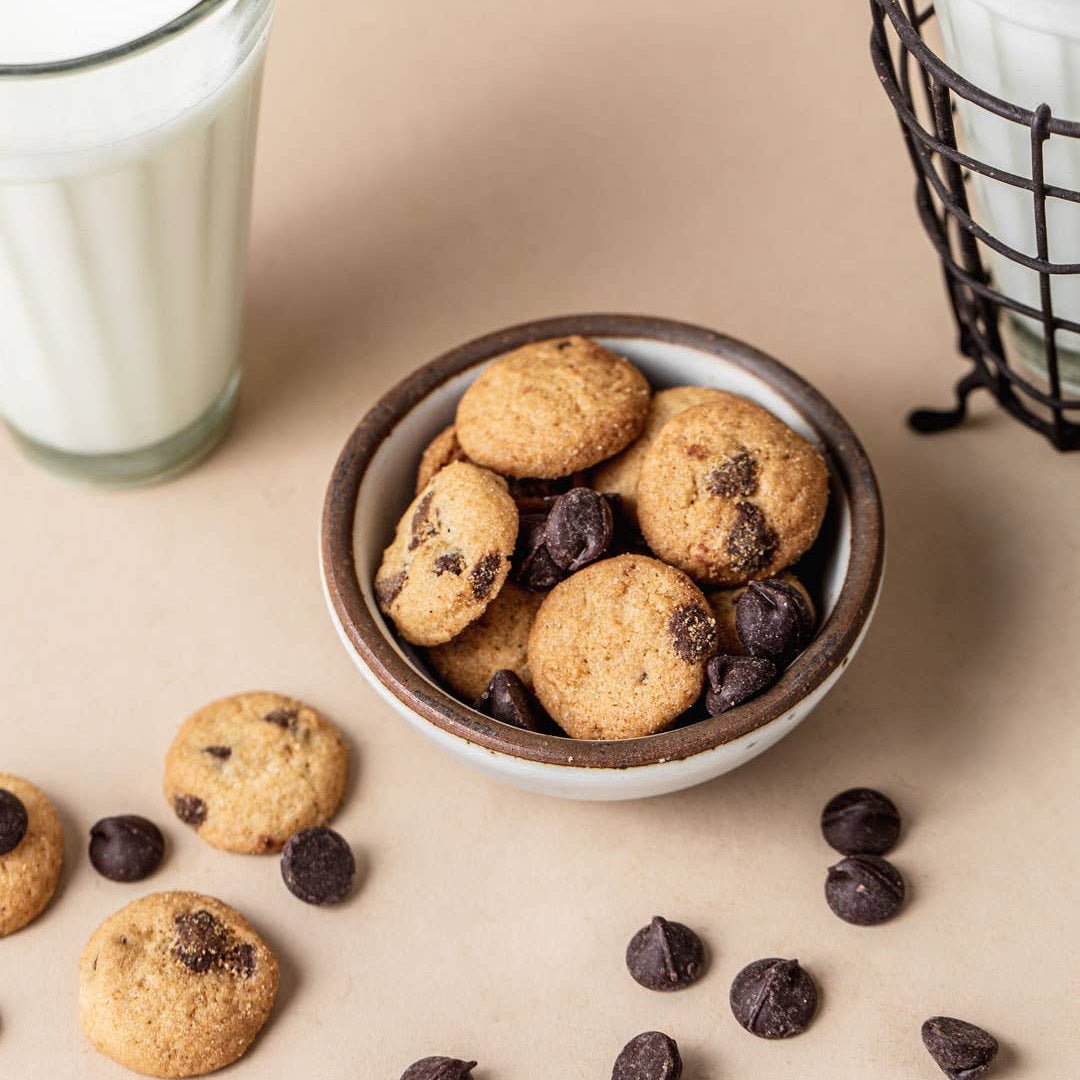 Cookies with chocolate chips in a bowl on a beige surface with a glass of milk.