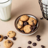 Cookies with chocolate chips in a bowl on a beige surface with a glass of milk.